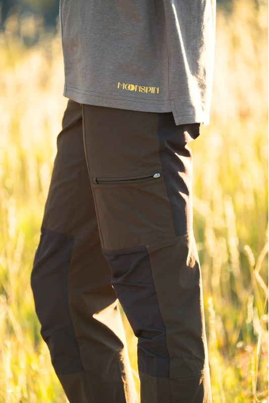 Woman wearing dark brown pants and a gray short with 'MOONSPIN' branding in a blurred natural setting.