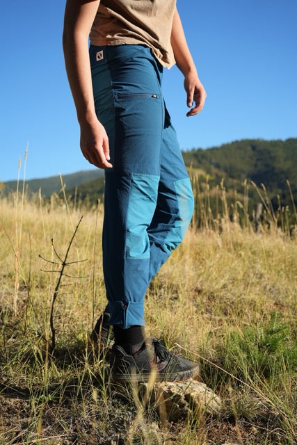 A woman outdoors in a grassy field wearing blue hiking pants, a tan top, and hiking shoes, with hills and blue sky in the background.